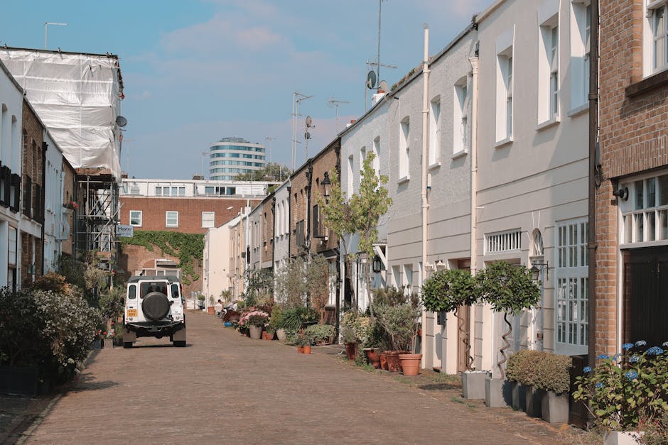 A narrow mews street with closely packed terraced houses on both sides, featuring brick and white-painted facades. The cobblestone pathway is lined with potted plants and small, well-maintained trees,