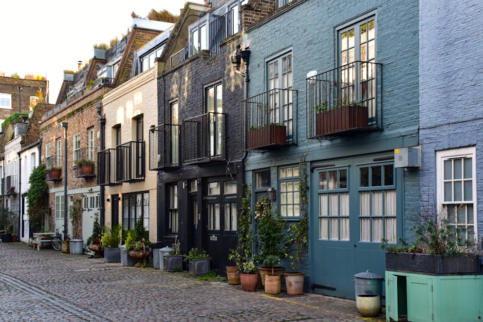 A narrow mews street with closely packed terraced houses on both sides, featuring brick and white-painted facades. The cobblestone pathway is lined with potted plants and small, well-maintained trees,