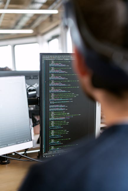 A close-up view of a laptop screen displaying lines of code in a programming editor, with a dark keyboard illuminated by soft pink and blue lighting. The screen shows a variety of code snippets, inclu