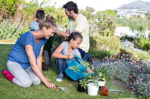 A family of four working together in a well-maintained garden in Bermondsey, with a lush green lawn, flower beds bordered by a variety of colourful flowering plants, and some gardening tools and pots placed on the grass. The foreground shows a woman in a blue top planting or tending to a small shrub, while a young girl in a blue jacket holds a watering can nearby. In the background, a man and a boy are engaged in garden tasks, with trees and bushes providing a natural backdrop. The garden is bathed in natural daylight, suggesting a clear, pleasant day, and the space appears to be part of a residential outdoor area suitable for family gardening activities, complementing local gardening services offered by Garden Maintenance Bermondsey.